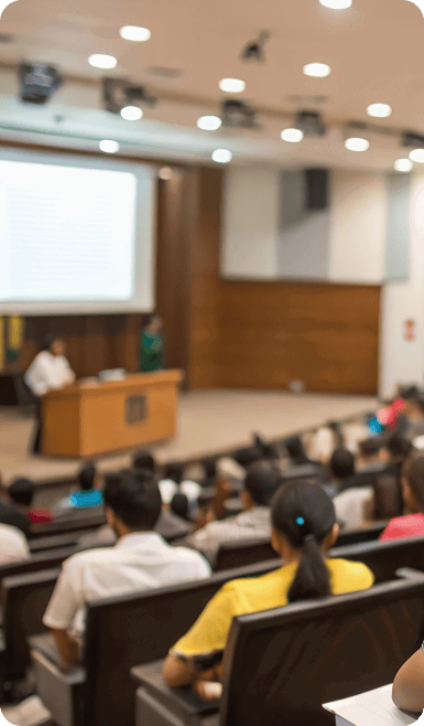 Students seated in an auditorium facing a stage with speakers and a large screen