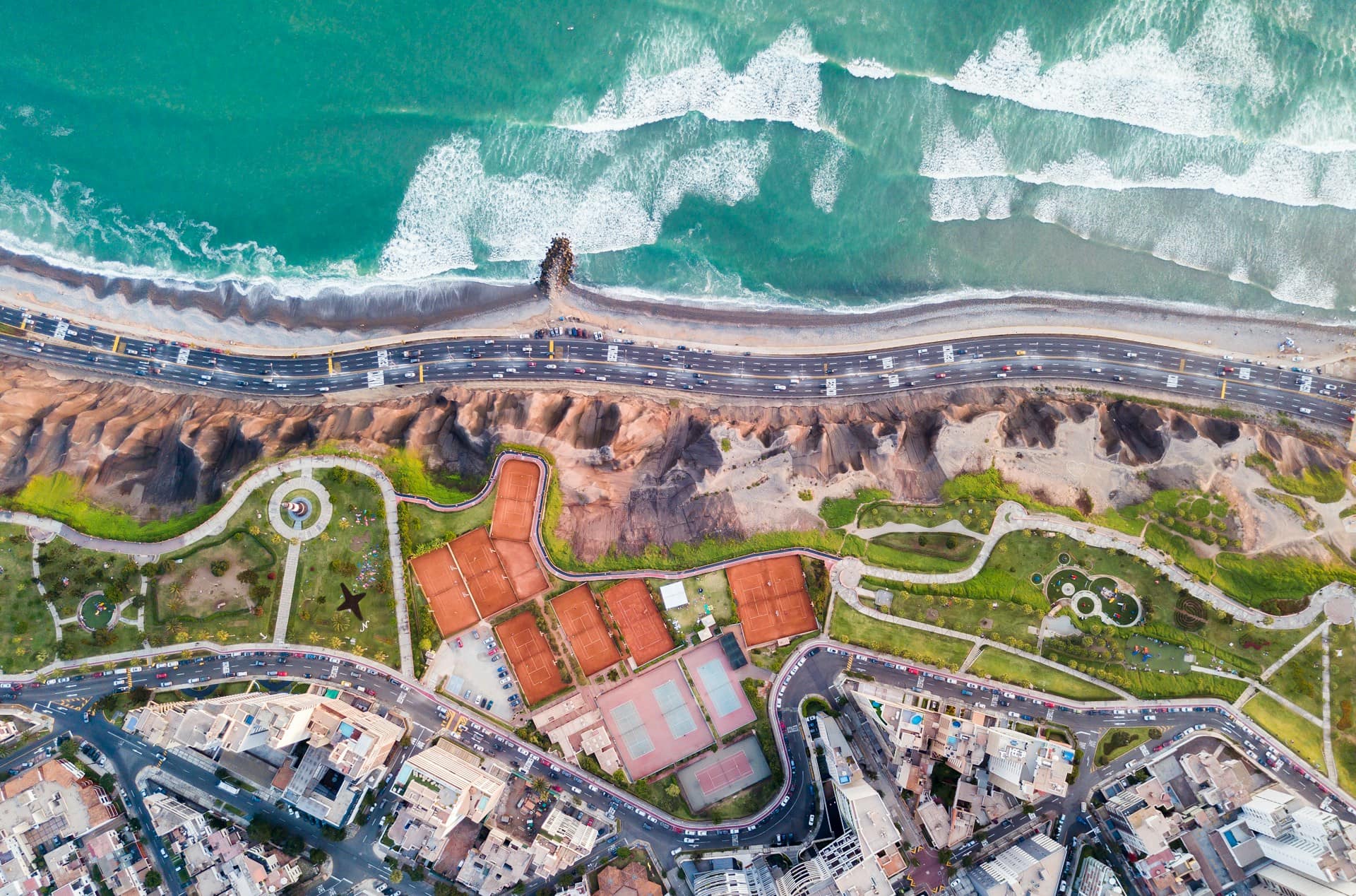 Aerial view of a coastal city with waves breaking on the shore, parallel roads running beside cliffs, green parks, and surrounding urban buildings.