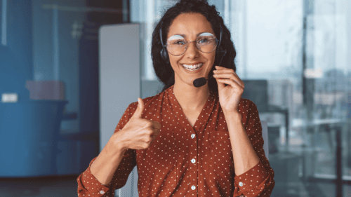 Smiling woman wearing glasses and a headset gives a thumbs-up gesture while sitting in an office setting, showcasing bilingual talent and suggesting positive customer service or support.