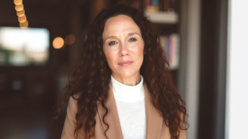 Erin Pedrami, a woman with long curly brown hair, wears a beige blazer over a white turtleneck as she stands indoors in soft lighting, shelves and blurred lights behind her. She has a neutral expression.